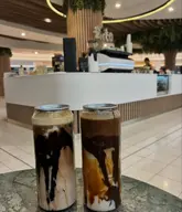 A photo of two canned iced lattes placed on a marble-effect table inside a shopping centre cafe. The drinks feature visible layers of chocolate sauce and coffee, presented in tall, clear cans.
