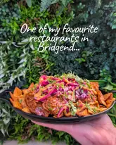 A close-up, handheld shot of a bowl of loaded nachos or a similar Mexican-inspired dish, presented against a green foliage wall. The image includes a text overlay promoting a restaurant in Bridgend.