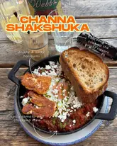 A bright, overhead shot of a Channa Shakshuka dish served in a cast-iron skillet, accompanied by a slice of toasted sourdough bread, set on a rustic wooden table.