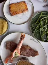 A high-quality, overhead flat-lay shot of a dinner spread at a brasserie, featuring a steak with a prawn, a side of mashed potatoes, green beans, and a potato dish. The lighting is bright and even, highlighting the textures of the food, and the composition is clean and professional.