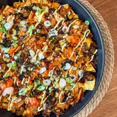 A top-down, high-quality food photography shot of a plate of Korean-fusion chicken nachos. The dish is generously topped with kimchi, spring onions, seaweed, and a drizzle of mayonnaise, presented on a dark plate over a textured placemat.