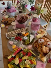 A high-angle, overhead shot of a lavish grazing table spread, featuring a variety of pastries, scones, cheeses, and fresh fruit, decorated with flowers and candles for a hen party celebration.