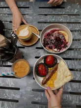 An overhead flat-lay shot of a brunch spread featuring pancakes with berry compote, a cooked breakfast plate with sausage, mushrooms, tomatoes, and flatbread, and a latte.