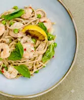 A clean, well-composed overhead shot of a prawn and pea pasta dish. The lighting is soft and natural, effectively highlighting the textures of the pasta and the freshness of the basil and lemon. The composition is balanced and centered, ensuring the dish is the clear focus without any distracting elements.
