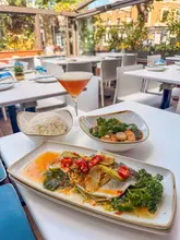 A bright, natural-light shot of a pan-Asian meal featuring a steamed fish dish, a side of rice, a small bowl of prawns, and a cocktail, set on an outdoor terrace.