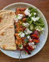 A high-quality, top-down food photography shot of a homemade chicken skewer meal. The plate features grilled chicken skewers topped with tzatziki and fresh herbs, served alongside warm flatbreads and a fresh green salad with grilled vegetables. The lighting is bright and even, highlighting the textures of the food.