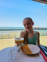 A young girl poses with a mocktail at a table in a restaurant with a view of the sea and pier in Blackpool.