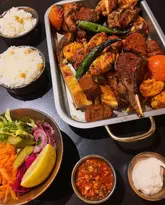 A top-down, high-angle shot of a Turkish mixed grill platter served on a metal tray, accompanied by two bowls of rice, a bowl of salad, and two small bowls of dips, all set on a dark table.