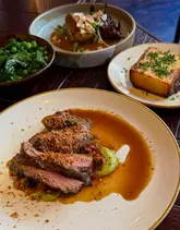 A high-quality, top-down shot of a spread of dishes including BBQ lamb rump, smoked potato gratin, and buttered greens, showcasing an elevated dining experience.