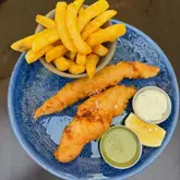 A top-down, natural light photograph of a fish and chips meal served on a blue plate. The dish consists of two pieces of battered haddock, a bowl of chunky chips, a small pot of mushy peas, and a small pot of tartar sauce, accompanied by a lemon wedge. The setting appears to be an outdoor table.