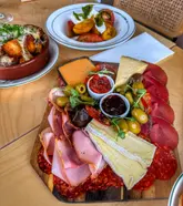 A high-quality, well-lit overhead shot of a charcuterie board featuring various meats, cheeses, olives, and dips, accompanied by a side of roasted potatoes and a tomato salad. The composition is clean and inviting, effectively showcasing the freshness and variety of the food, making it a strong piece of visual marketing for a lunch venue.