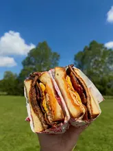 A bright, high-quality close-up shot of a breakfast sandwich held against a clear blue sky and green field background. The natural lighting is excellent, effectively highlighting the textures of the toasted bread, sausage, bacon, and egg.