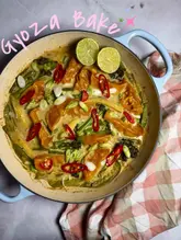 A vibrant, overhead shot of a Thai green curry gyoza bake served in a blue enamel pan. The dish is garnished with fresh red chili slices, spring onions, and lime halves, set against a marble background with a checkered cloth.