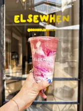 A vibrant, well-composed product shot of a smoothie held in front of the 'Elsewhen' shopfront, effectively using the brand's signage as a backdrop to establish the venue.