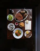 A high-quality, overhead flat-lay shot of a table spread at a restaurant, featuring a variety of dishes including duck, salmon, oysters, and fries, alongside a menu. The lighting is moody and atmospheric, highlighting the textures of the food.
