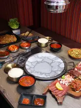 A top-down, overhead shot of a Korean BBQ table spread, featuring a central grill, raw meat platters, bowls of rice, kimchi, and various dipping sauces, capturing the communal dining experience.