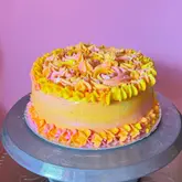 A vibrant, multi-colored vanilla sponge cake with strawberry jam filling, displayed on a metal cake turntable against a plain pink background.