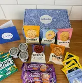 A flat-lay photograph of a 'Nibula' treat box, featuring various packaged biscuits, jams, hot chocolate sachets, and tea bags spread out on a kitchen counter.
