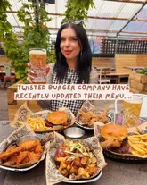 A bright, casual, and inviting shot featuring a creator holding a pint of beer at a table filled with burgers, loaded fries, and chicken tenders. The setting appears to be a covered outdoor area of a brewery/restaurant, with the food presented in branded paper baskets. The image effectively communicates a relaxed, social dining experience.