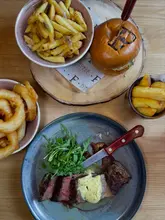 A high-angle flat-lay shot of a meal spread featuring a flat iron steak with garlic herb butter, a burger, a bowl of fries, and a bowl of onion rings on a wooden table.