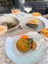 A flat-lay photograph of a three-course meal spread on an outdoor table at Crowded House, featuring cocktails, a main dish, and sides.