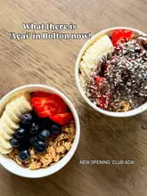 A high-angle, well-lit photograph of two acai bowls served in paper containers on a wooden table. The bowls are topped with fresh fruit, granola, and in one instance, Nutella and coconut flakes, presenting a clean and appetizing aesthetic.