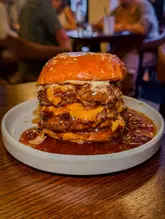 A close-up, eye-level shot of a double-patty cheeseburger with a significant amount of sauce, served on a white plate on a wooden table. The background shows a blurred restaurant setting with other diners.