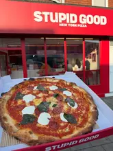 A casual, first-person perspective shot of a pizza box held in front of a restaurant storefront. The image features a pepperoni and burrata pizza, showcasing the crust and toppings, with the venue's branding visible in the background.