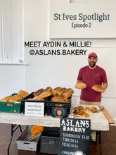 A promotional image for a local artisan bakery, featuring the owner holding two loaves of sourdough at a market stall. The image includes text overlays introducing the business and the owners.