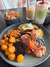 A high-quality, well-lit photograph of a hearty full English breakfast served alongside a side of tater tots and a vibrant matcha latte. The image captures the textures of the cooked breakfast items effectively, using natural light to highlight the freshness of the poached egg and the crispiness of the bacon.