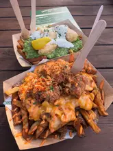 A high-angle, natural light shot of two loaded chip dishes served in cardboard trays on a wooden table. The image clearly showcases the textures and toppings of the halloumi and mac & chicken loaded chips, effectively communicating the casual, indulgent nature of the street food offering.