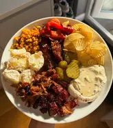 A home-cooked 'picky bits' dinner plate featuring various pork items, potato salad, grilled vegetables, and a whipped feta dip.