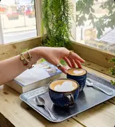 A high-quality, aesthetically pleasing shot of two coffees served on a metal tray next to a book, captured in a bright, natural-light setting. The composition is balanced, featuring a hand reaching for a cup, which adds a human touch and a sense of warmth.