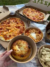 A top-down, flat-lay style photograph of a takeaway picnic spread on a blanket, featuring two pizzas, a garlic and mozzarella bombe, a bowl of spaghetti, and a bowl of calamari. The lighting is natural and warm, consistent with an outdoor sunset setting.