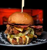 A close-up, high-contrast shot of a fried chicken burger featuring bacon, cheese, and pickles, set against a dark background. The lighting is punchy and direct, highlighting the textures of the fried chicken and the sauce drip.