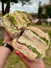 A bright, outdoor shot featuring four halves of generously filled focaccia sandwiches held by two people. The focus is sharp on the textures of the bread and fillings, making the food look fresh and appealing.