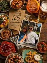 A high-quality, overhead flat-lay photograph of a birthday celebration at a tapas restaurant. The table is filled with a variety of dishes including shrimp, gnocchi, meatballs, octopus, calamari, and a tomato salad, accompanied by an Aperol Spritz and a beer. A birthday card and a photograph are placed centrally, adding a personal and celebratory atmosphere to the scene.