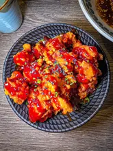 A vibrant, top-down close-up shot of Korean fried chicken glazed in a glossy red sauce, garnished with black sesame seeds and chopped spring onions, served in a dark, patterned bowl on a wooden table.