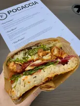 A high-quality, close-up shot of a 'poccacia' sandwich held in hand, effectively showcasing the cross-section and layered ingredients. The background features the venue's menu, which is softly blurred to provide context without distracting from the main subject.