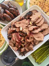 A top-down, natural light photograph of a home-cooked BBQ spread featuring sliced lamb, sausages, flatbreads, and asparagus.