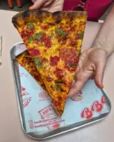 A high-quality, close-up shot of a large, New York-style pizza slice being held up, showcasing the texture of the cheese and sauce. The branding on the serving tray is clearly visible, adding context to the venue.