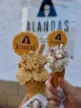 A bright, handheld shot of two gelato cones held up against a wall featuring the Alandas brand logo. The focus is sharp on the ice cream, which looks fresh and well-scooped.