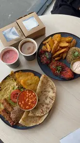 A top-down, casual shot of a Desi breakfast spread featuring grilled chicken, wedges, rice, and various teas, served on a mix of plates and takeaway packaging.