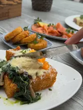 A close-up, action-oriented shot of a breakfast dish being cut with a knife, featuring a crispy hash brown base topped with sautéed spinach, mushrooms, and a poached egg, with additional breakfast items like smoked salmon visible in the background.