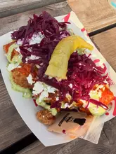 A close-up, high-angle shot of a portion of chicken wings topped with red cabbage, feta cheese, sauce, and a whole pickled pepper, served in a paper tray on a wooden table.