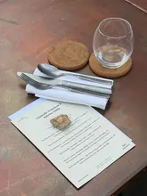 A clean, professional flat-lay photograph of a tasting menu at the Carousel x Ashcombe Estate pop-up, featuring elegant cutlery and glassware on a rustic table surface. The image focuses on the menu details, conveying an atmosphere of high-end, exclusive dining.