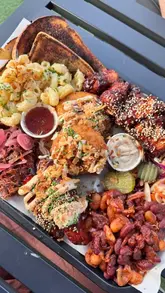 A promotional video for Fat Jack's Shack in Chichester, featuring close-up shots of a large BBQ sharing platter, including pulled pork, mac and cheese, wings, and a savory donut. The video also provides a brief tour of the venue's outdoor seating area at Draper's Yard.