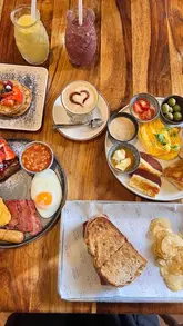 A high-angle, flat-lay shot of a brunch spread featuring pancakes, a full English-style breakfast, a sandwich with chips, and various drinks on a wooden table.