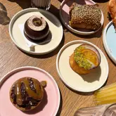 A flat-lay photograph of a selection of pastries and desserts from a bakery, including a chocolate mirror gateaux, a sausage roll, a cheese straw, and an orange cake, arranged on a wooden table.