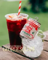 A bright, outdoor promotional shot featuring a plastic cup of sangria and a canned margarita served over ice, captured at the Edinburgh Food & Folk Festival in George Square. The image effectively communicates the casual, summery atmosphere of the event.